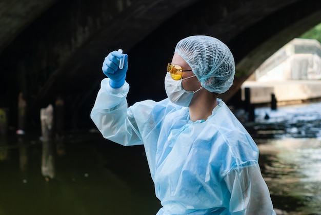 Female ecologist or epidemiologist takes testtube water analysis in city river under an old bridge