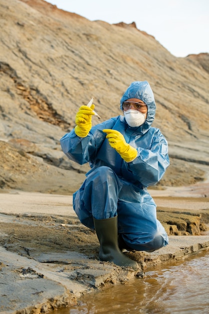 Photo female ecologist in gloves, coveralls, respirator and eyeglasses looking at flask with sample of dirty water or soil in dangerously polluted area