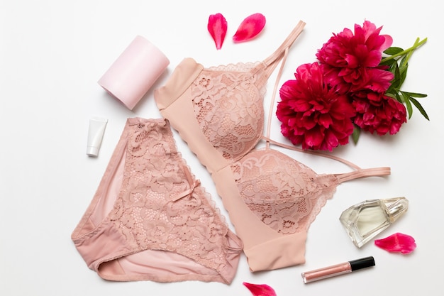 Female elegant pink lace bra, flat peony, lipstick, hand cream and barrette on white 