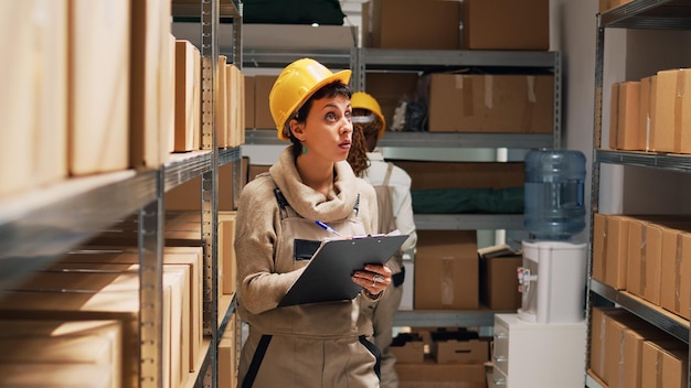 Female employee checking list of goods on papers, using files to do cargo inventory and logistics. Woman in overalls counting number of cardboard boxes or packages in storage room.