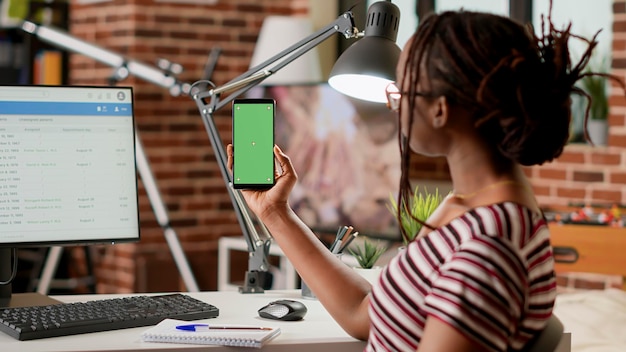 Female employee holding smartphone with greenscreen display at\
home, working remotely. woman looking at mobile phone with isolated\
chroma key, blank copyspace template and mockup background.