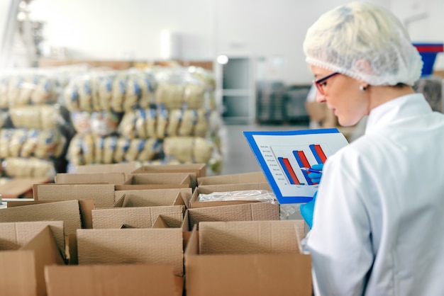 Foto impiegata femminile in uniforme sterile e con il grafico in mano contando i prodotti in scatola interno di fabbrica alimentare.