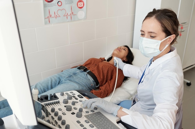 Женщина-эндокринолог в белом халате и маске сидит у ультразвукового оборудования