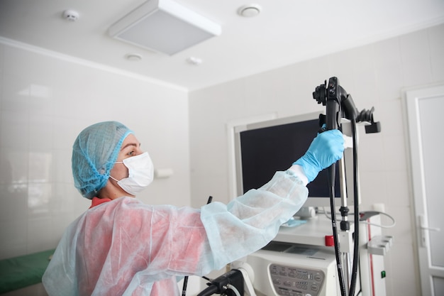 A female endoscopist surgeon in a protective suit, cap, mask and gloves picks up an endoscope before starting procedure