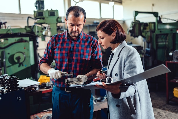 Female engineer discussing with machinist holding cogwheel and analyzing blueprint