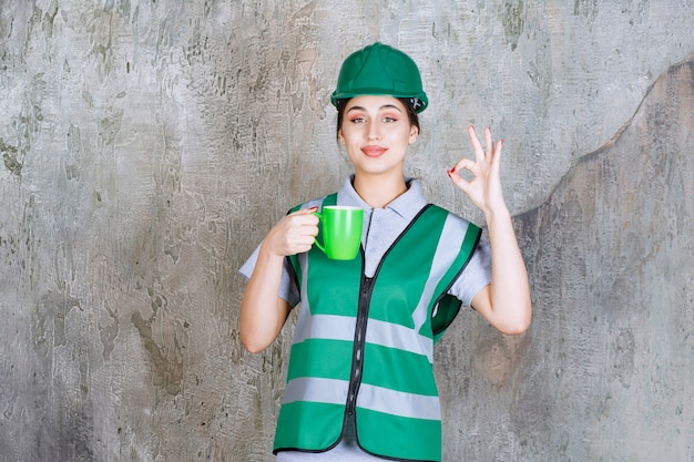 緑のコーヒーマグカップを保持し、楽しみのサインを示す緑のヘルメットの女性エンジニア