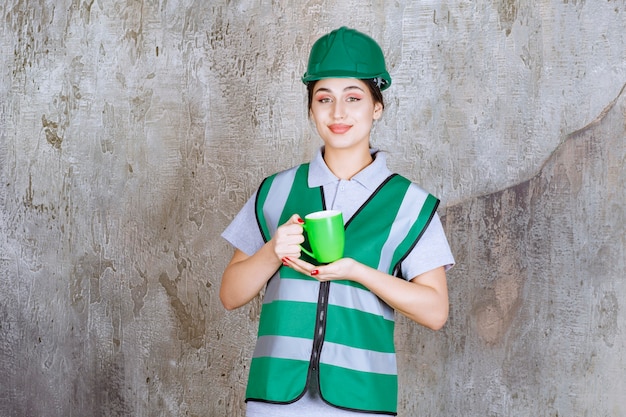 Female engineer in green helmet holding a green coffee mug. 