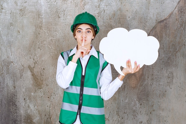 Female engineer in green uniform and helmet holding a cloud shape info board and asking for silence