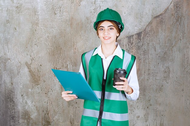 Female engineer in green uniform and helmet holding a green project folder and a cup of drink