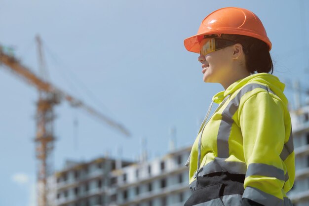 A female engineer in a hard hat and goggles on the background of a multi-storey building under construction.
