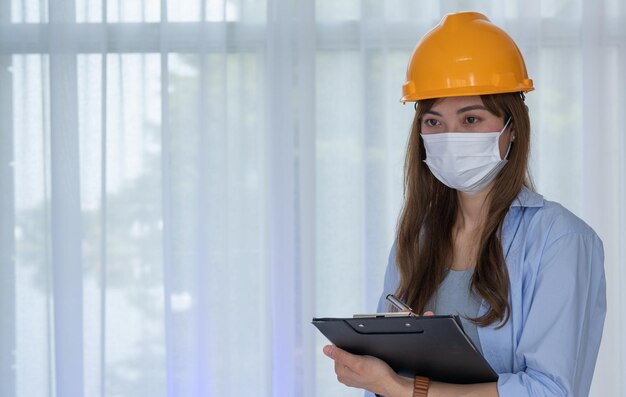 L'ingegnere femminile indossa una maschera facciale con un casco giallo di sicurezza controlla la correttezza della costruzione