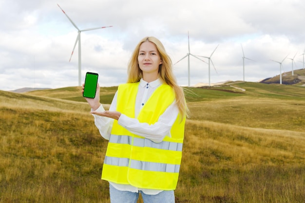 風力タービンの背景に携帯電話を持つ女性エンジニア 緑の画面を持つスマートフォン 環境に優しいエネルギー