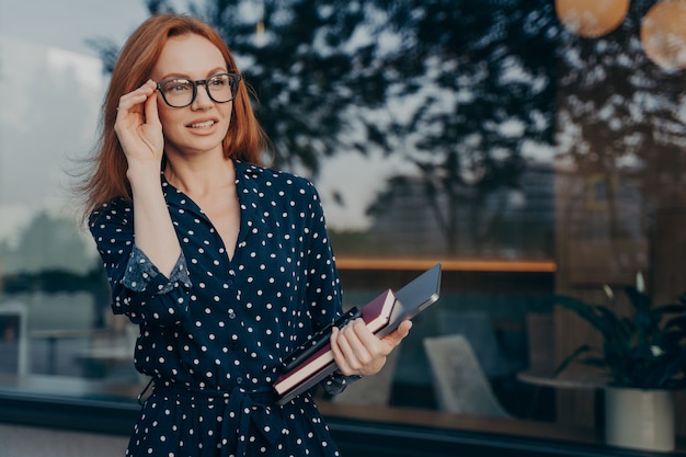 Female entrepreneur carries tablet smartphone and organizer poses near window of restaurant