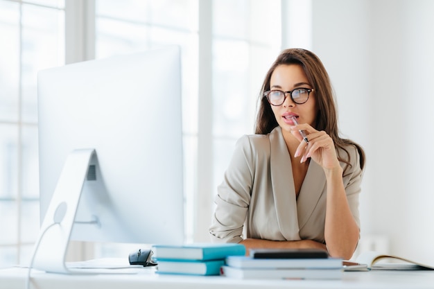 Female entrepreneur keeps pen in mouth, focused in monitor of computer