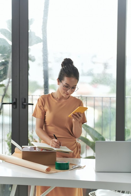 Female entrepreneur preparing parcel boxes and packing goods while checking orders on her mobile