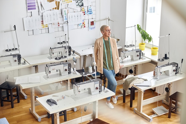 Female entrepreneur in stylish outfit with tape measure leans onto table in empty sewing workshop 