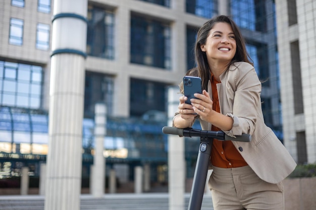 Female entrepreneur with smartphone while driving an electrical push scooter near business center on city street Transportation environmental concept