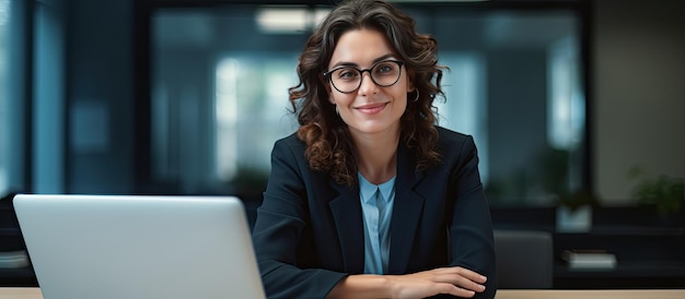 Female entrepreneur working at desk in office smiling at camera with empty laptop screen space for copy