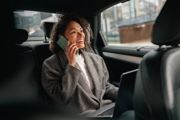 Female entrepreneur working on laptop and talking phone during traveling to office in luxury car