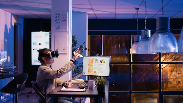 Female entrepreneur working at night with vr glasses, using virtual reality and 3d environment on headset. Business manager planning financial investment with interactive goggles. Handheld shot.