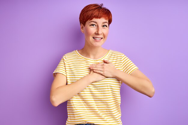 Female expressing gratitude, holding hands on chest, feeling kindness. isolated over purple wall