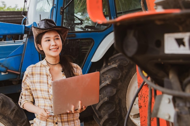 Female farmer cattle agribusiness operator operator holding laptop inspects machines and tractors.