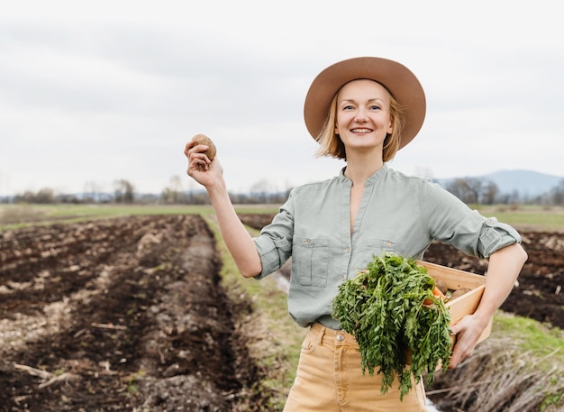 Female farmer holding wooden box full of fresh raw vegetables in agricultural field area