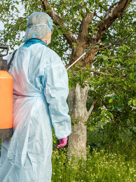 保護服を着た女性農家が、春の果樹園で真菌病や害虫のリンゴの木に圧力噴霧器と化学薬品を噴霧しています。