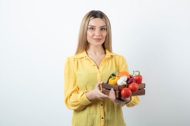 Female farmer standing and holding a wooden crate full of a variety of organic vegetables