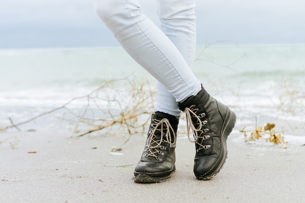 Female feet in blue jeans and black winter boots standing in the sand at the beach