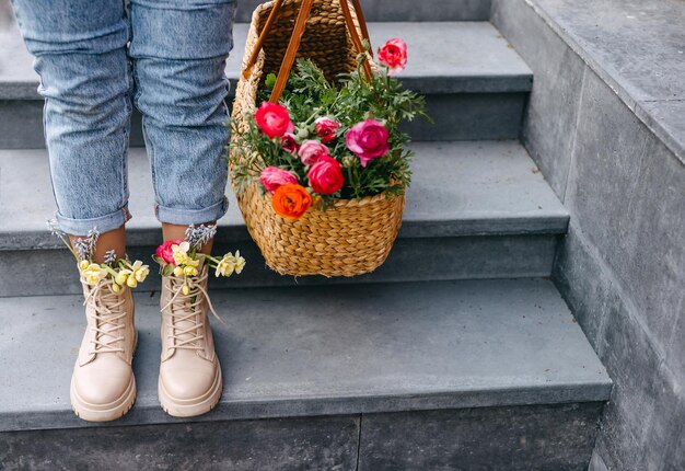 Female feet in boots with different colorful spring flowers inside next to a big basket with flowers