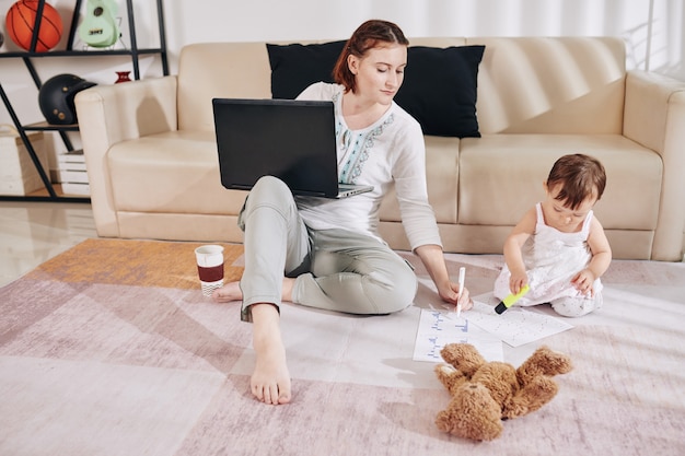 Female financial analyst in loungewear working from home, her little daughter sitting o ther floor nearby and playing with highlighter