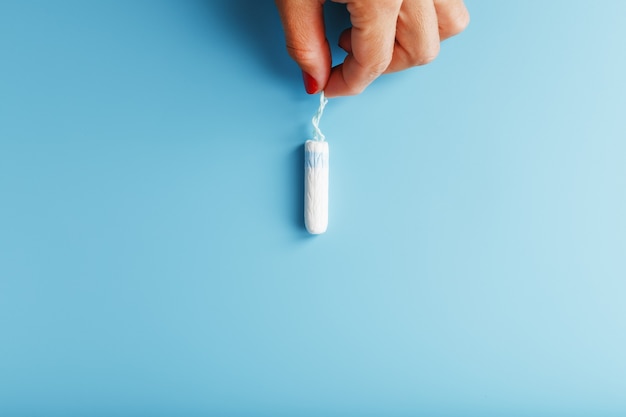 Female fingers with red nails hold a female sanitary swab on a blue background with a free space. Menstrual cycle