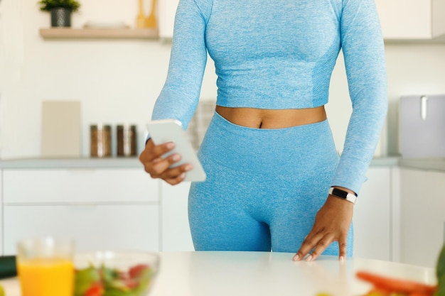 Female fitness blogger using smartphone in the kitchen black\
lady in sportswear preparing healthy dinner at home