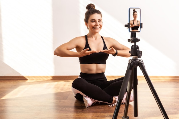 Female fitness instructor recording tutorial video of breathing technique after workout wearing black sports top and tights Full length studio shot illuminated by sunlight from window