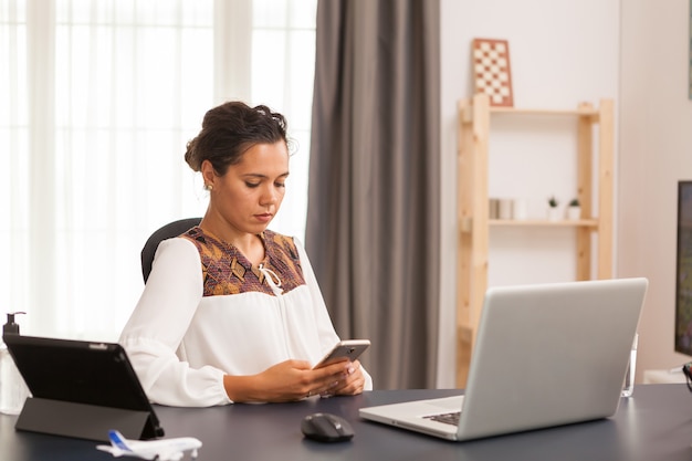 Female freelancer browsing on smartphone while working from home office.