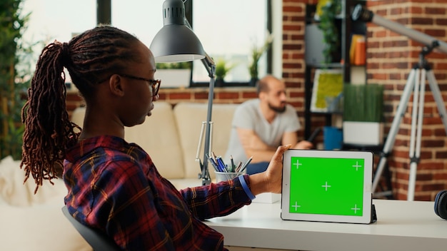 Female freelancer looking at digital tablet with horizontal greenscreen at home office desk, using chroma key display with green screen mockup template. Analyzing isolated copyspace on device.