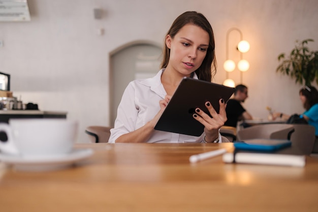 デジタル タブレットを使用してカフェで働く女性フリーランサーの若いビジネス マネージャーがコーヒーを飲む