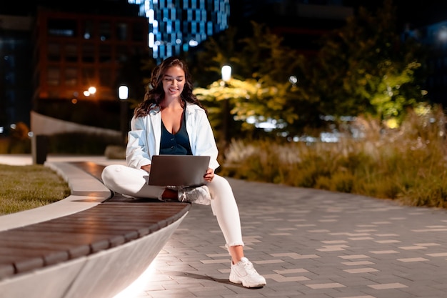 Female freelancer working on laptop sitting on bench at night in city park on modern urban