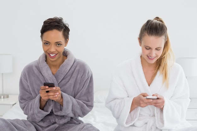 Female friends in bathrobes text messaging on bed