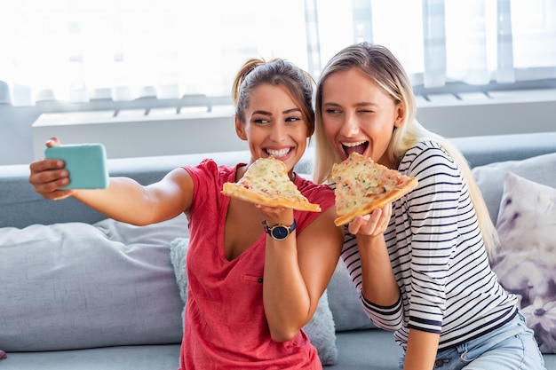 Female friends eating pizza with wine on sofa at home