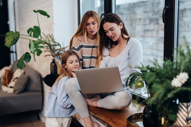 Female friends and a little girl sit together and look at the laptop screen.