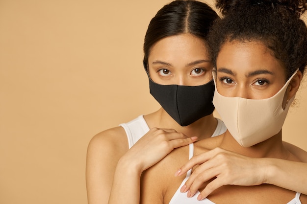 female friends wearing protective facial masks while posing together isolated over beige background.