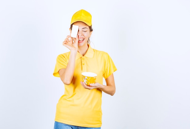 Ragazza femminile in uniforme gialla che consegna una tazza di noodle da asporto gialla e presenta il suo biglietto da visita al cliente.