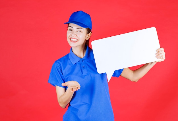 Женщина-гид в синей форме держит белую прямоугольную информационную доску и приглашает человека впереди поучаствовать.