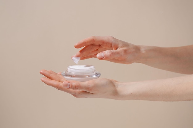 Female hand applying white moisturizing cream from glass jar on finger on beige isolated background Concept of beauty cosmetic product skin treatment and protection