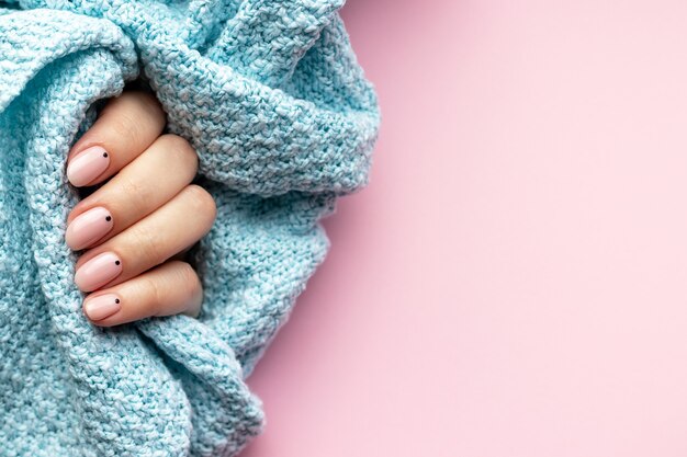 Mano femminile in un tessuto maglione lavorato a maglia blu con bella manicure alla moda - unghie nude rosa con piccoli punti neri su uno sfondo rosa con spazio di copia