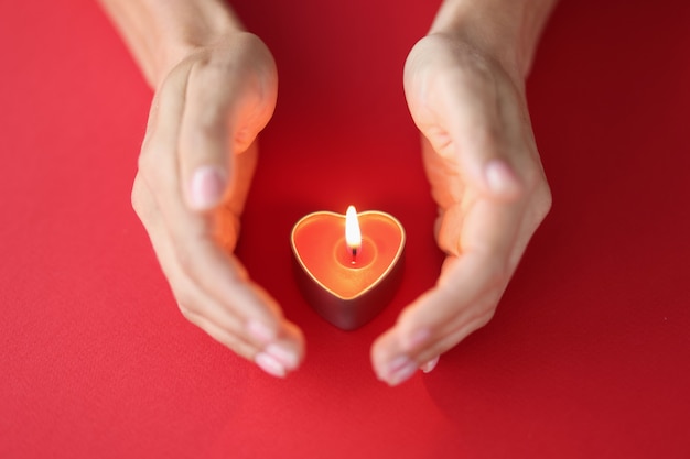 여성의 손은 빨간색 배경 촛불에 하트 모양으로 불타는 촛불을 조심스럽게 보호합니다.