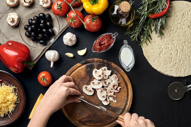 Female hand cut mushrooms on wooden board on kitchen table around lie ingredients for pizza vegetables cheese and spices