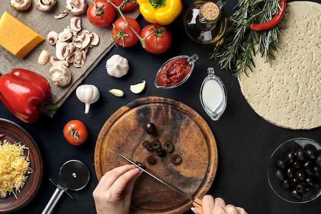Фото Женские оливки ручной работы на деревянной доске на кухонном столе вокруг лежат ингредиенты для пиццы, овощей, сыра и специй